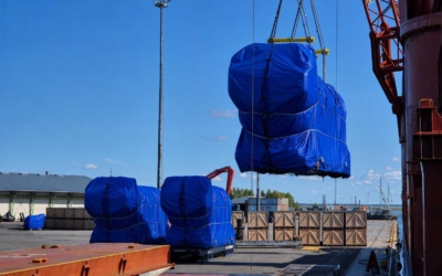 Breakbulk cargo units for a power plant project sit tarp-wrapped on the quay, one lifted mid-air by crane beside wooden shipping crates.