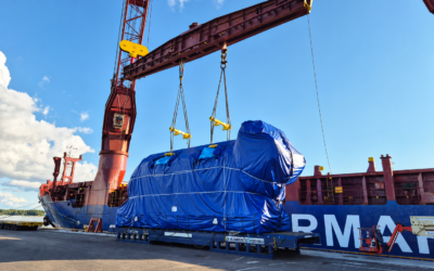 A tarp-wrapped generator unit swings from crane hooks onto a low-loader trailer during an oversized cargo transport operation.