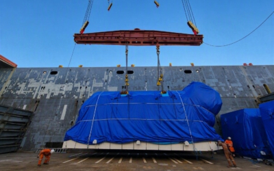 Power plant equipment lowered into a vessel's open cargo hold, guided by dock workers in orange overalls during breakbulk loading.