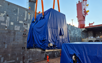 A blue tarp-wrapped engine unit descends into a ship's hold on lifting straps at dusk, part of a heavy-lift shipping operation.