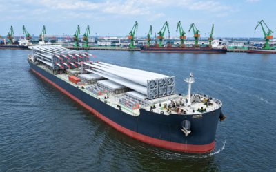 Bulk carrier loaded with wind turbine blades sails past a port lined with green gantry cranes, leaving a visible wake