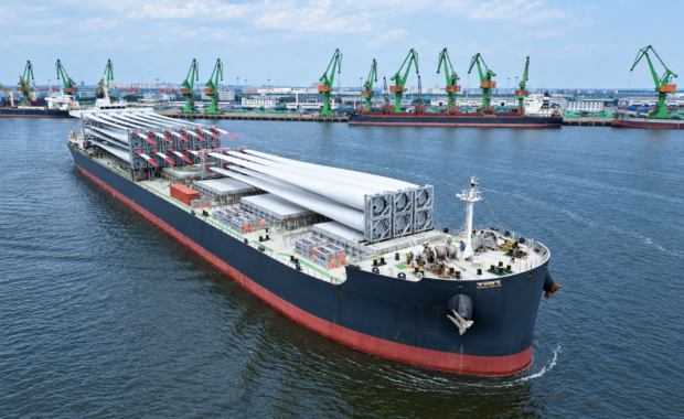 Bulk carrier loaded with wind turbine blades sails past a port lined with green gantry cranes, leaving a visible wake