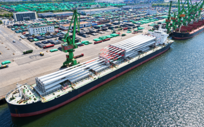 Aerial view of the moored blade-carrying vessel beside a cargo yard, with wind turbines visible on the horizon.