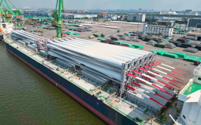 Ship docked at the quay with rows of wind turbine blades, red and white tips extending over stacked steel pipes on the pier.
