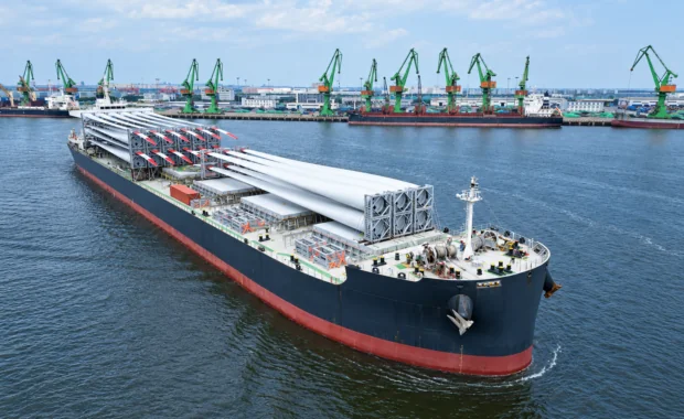 Bulk carrier loaded with wind turbine blades sails past a port lined with green gantry cranes, leaving a visible wake