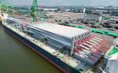 Ship docked at the quay with rows of wind turbine blades, red and white tips extending over stacked steel pipes on the pier.